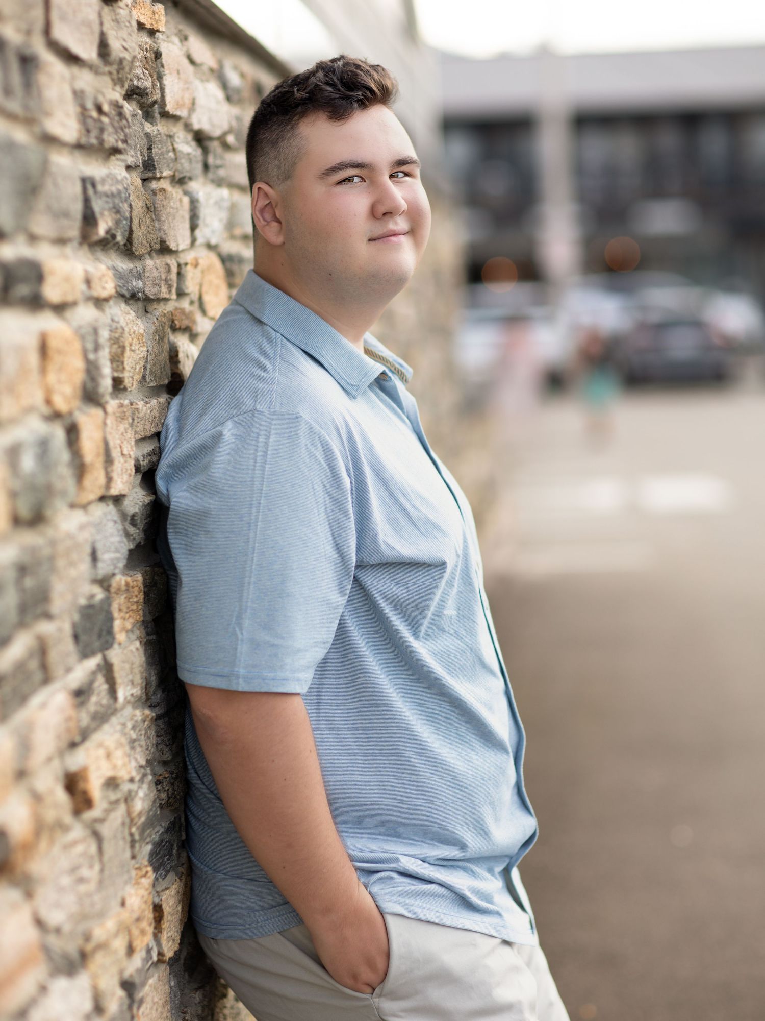 Young man in light blue polo shirt leaning against brick wall outdoors, smiling confidently.