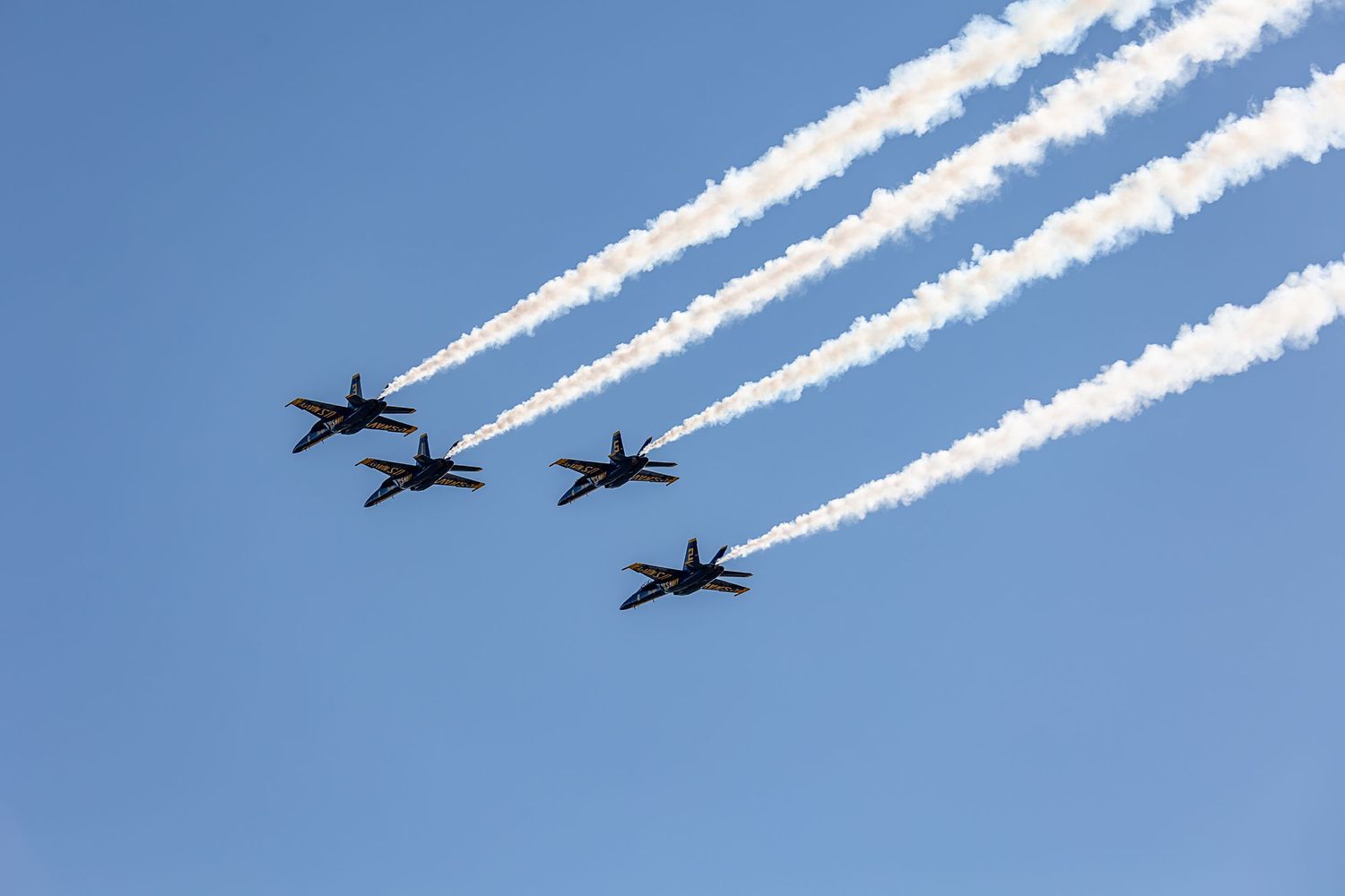 Military jets flying in formation leave white contrails against a bright blue sky during an aerial demonstration.