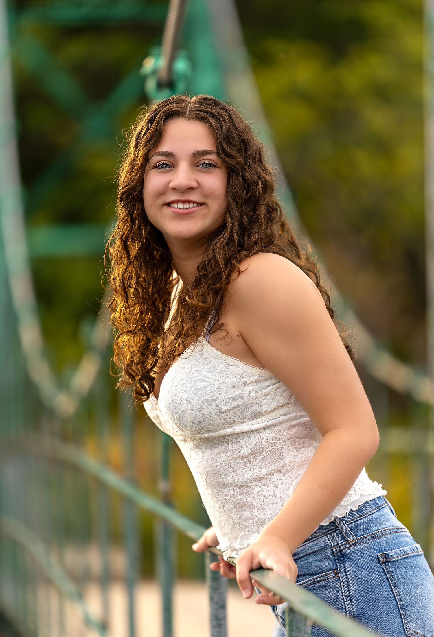 A graduating senior with long curly brown hair wearing a white top and jeans leans against a railing outdoors on a sunny day.