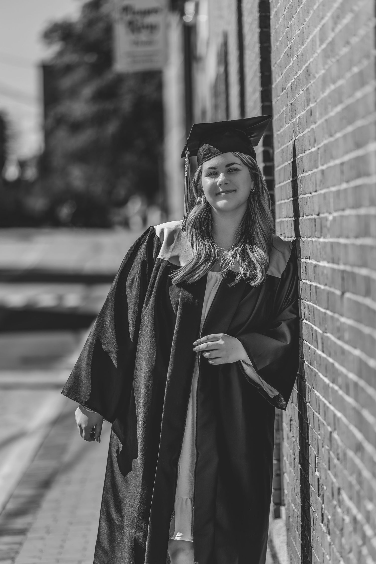 Black and white graduation portrait of a graduate in cap and gown leaning against a brick wall on a sunny day.
