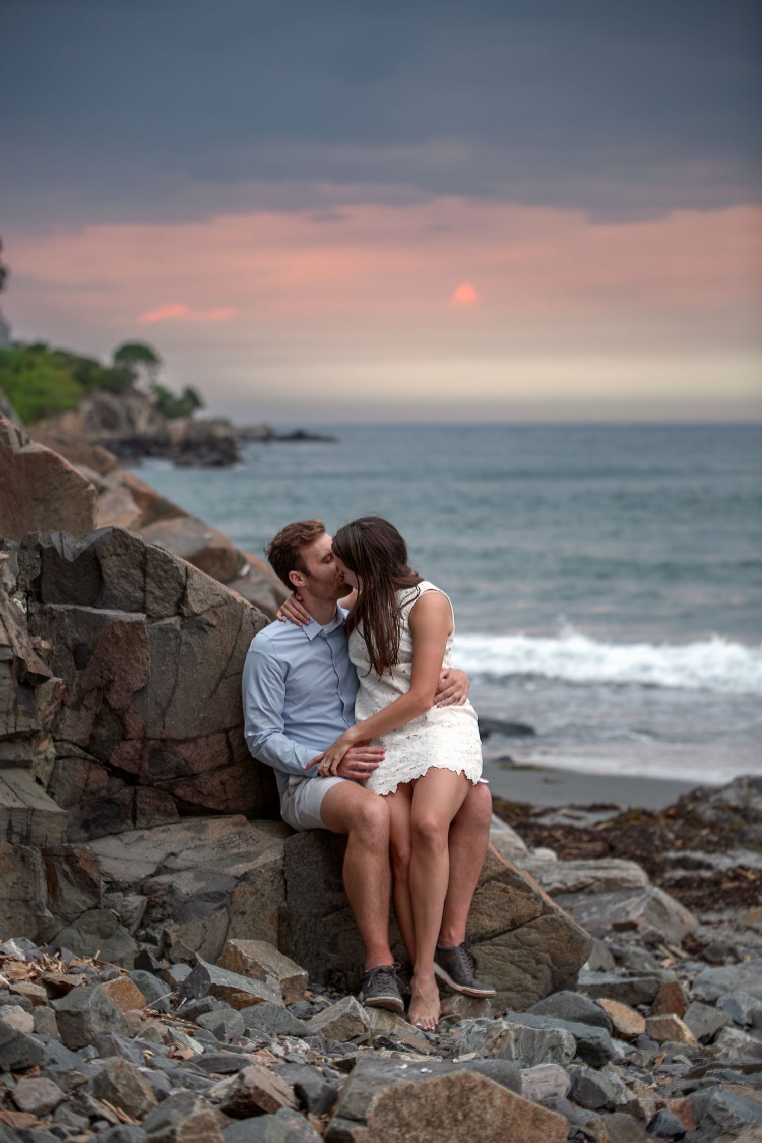 Couple sharing a romantic kiss on rocky beach at sunset with ocean waves and pink sky backdrop.