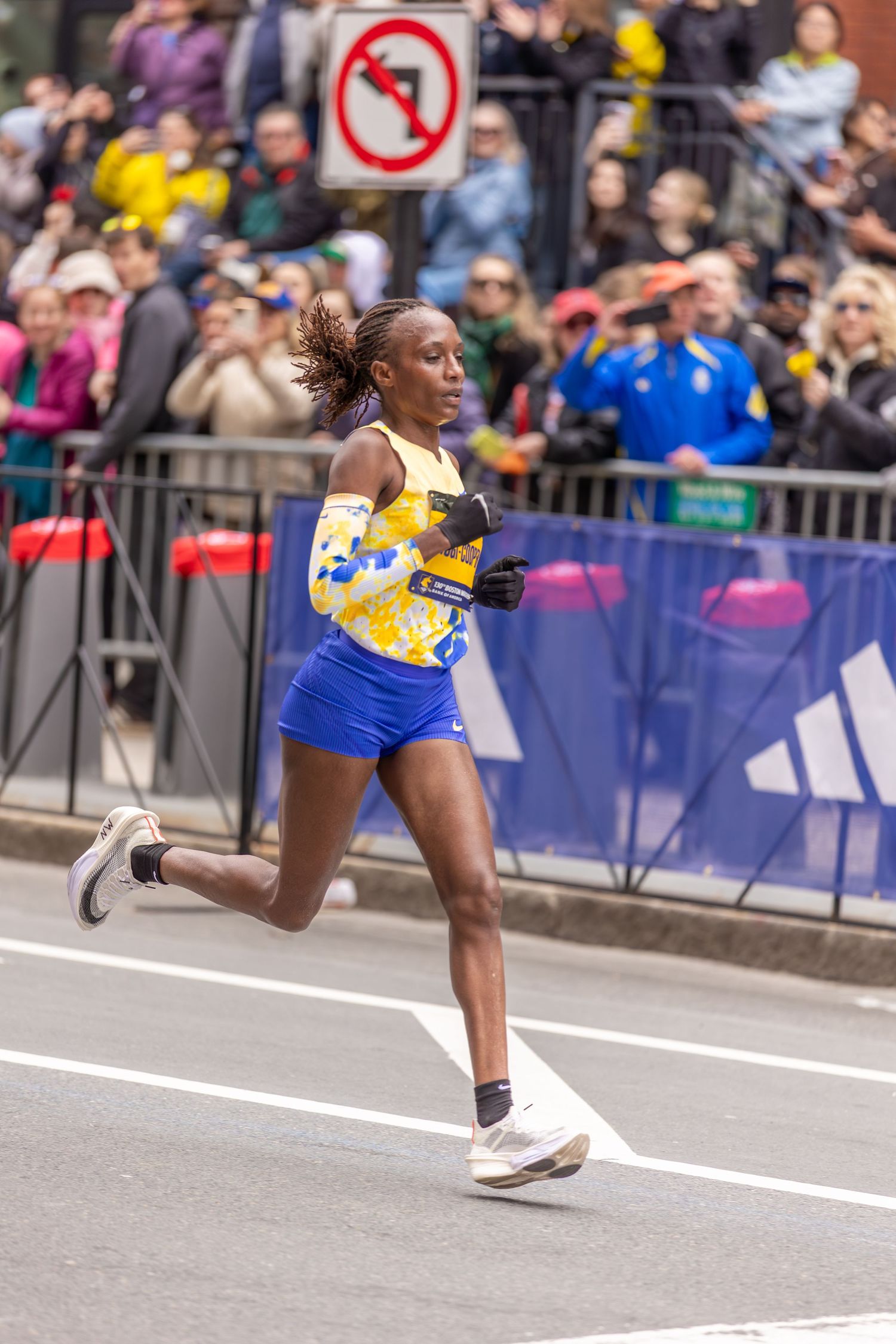 Female runner competing in Boston Marathon wearing blue shorts and yellow top, racing past crowds of spectators lining the street.