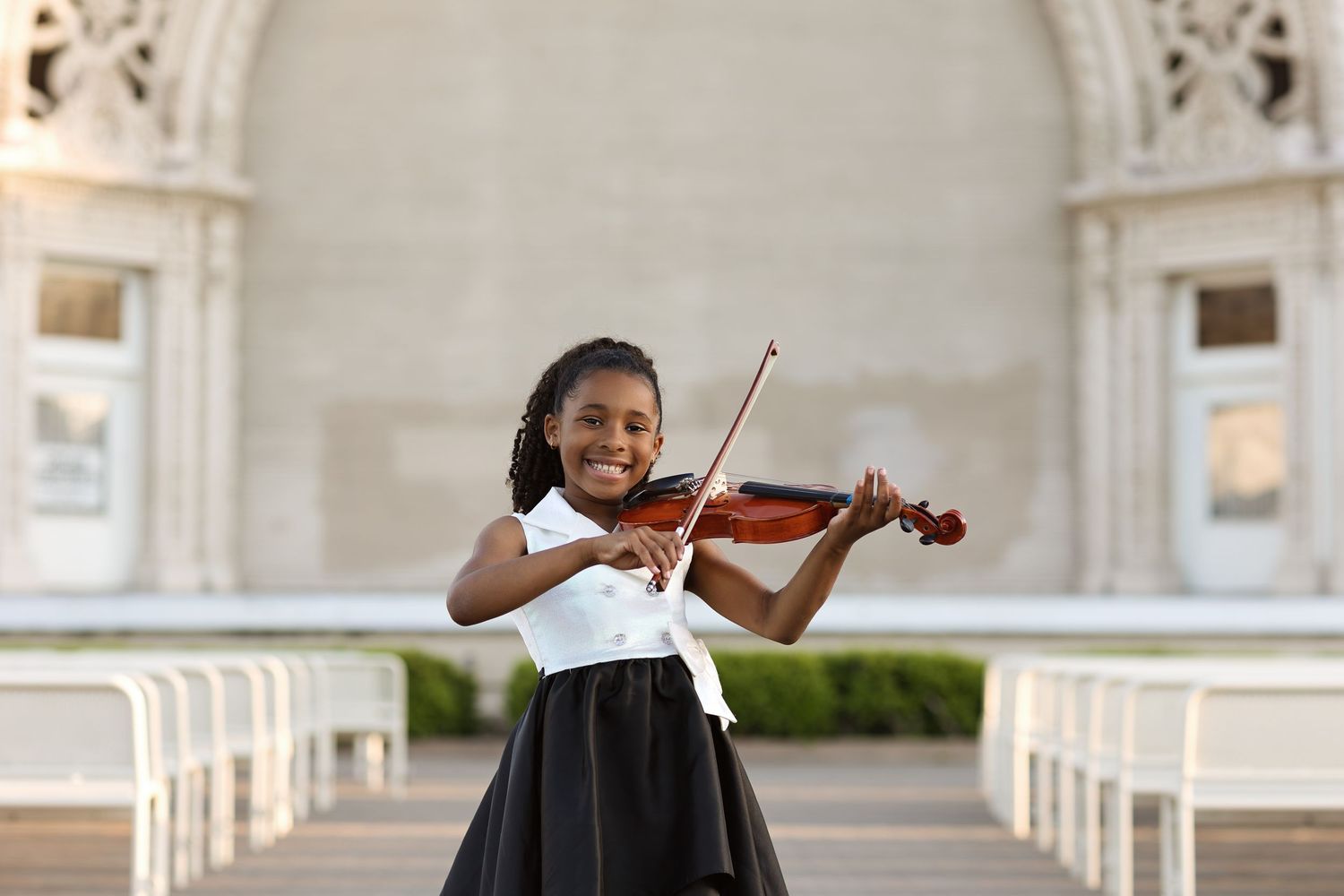 Young girl in black and white dress smiling while playing violin outdoors near elegant white columns.