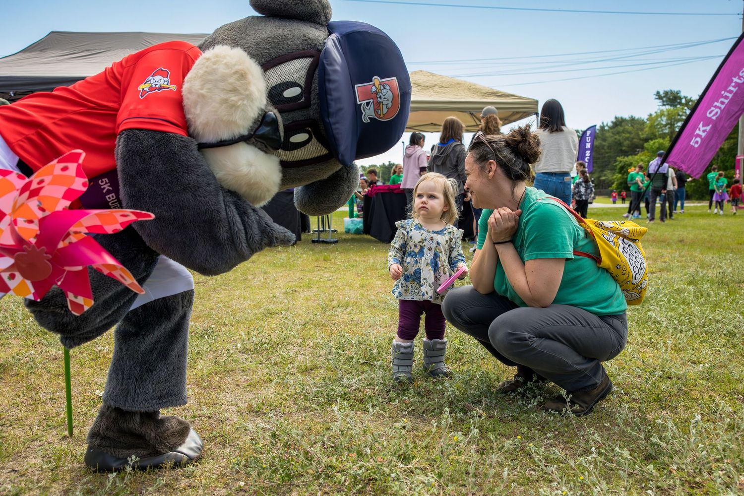 A sports mascot greets a toddler girl while a woman crouches nearby at an outdoor community event.