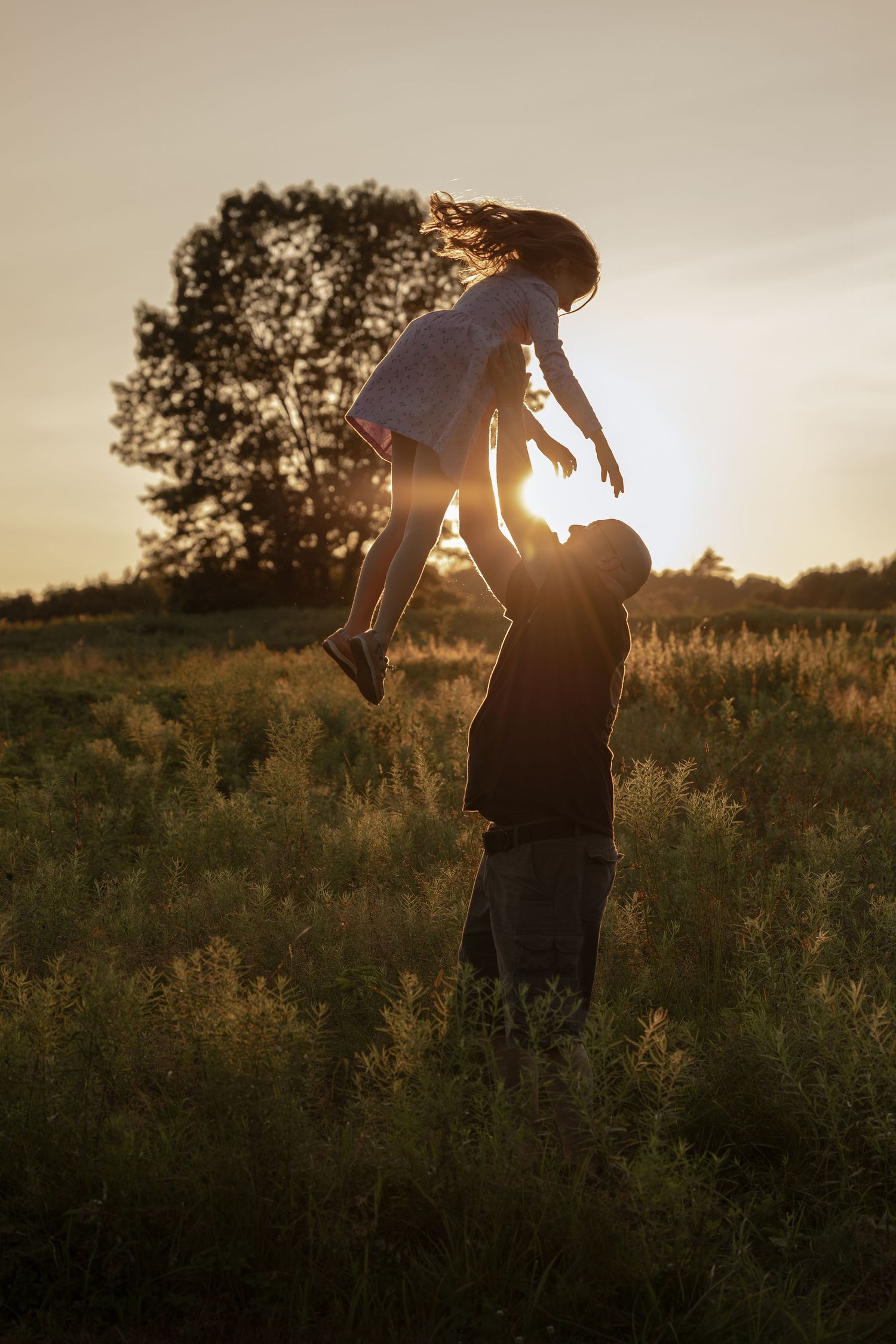 Father tossing his daughter in the air at sunset in a golden field, silhouetted against warm glowing light.