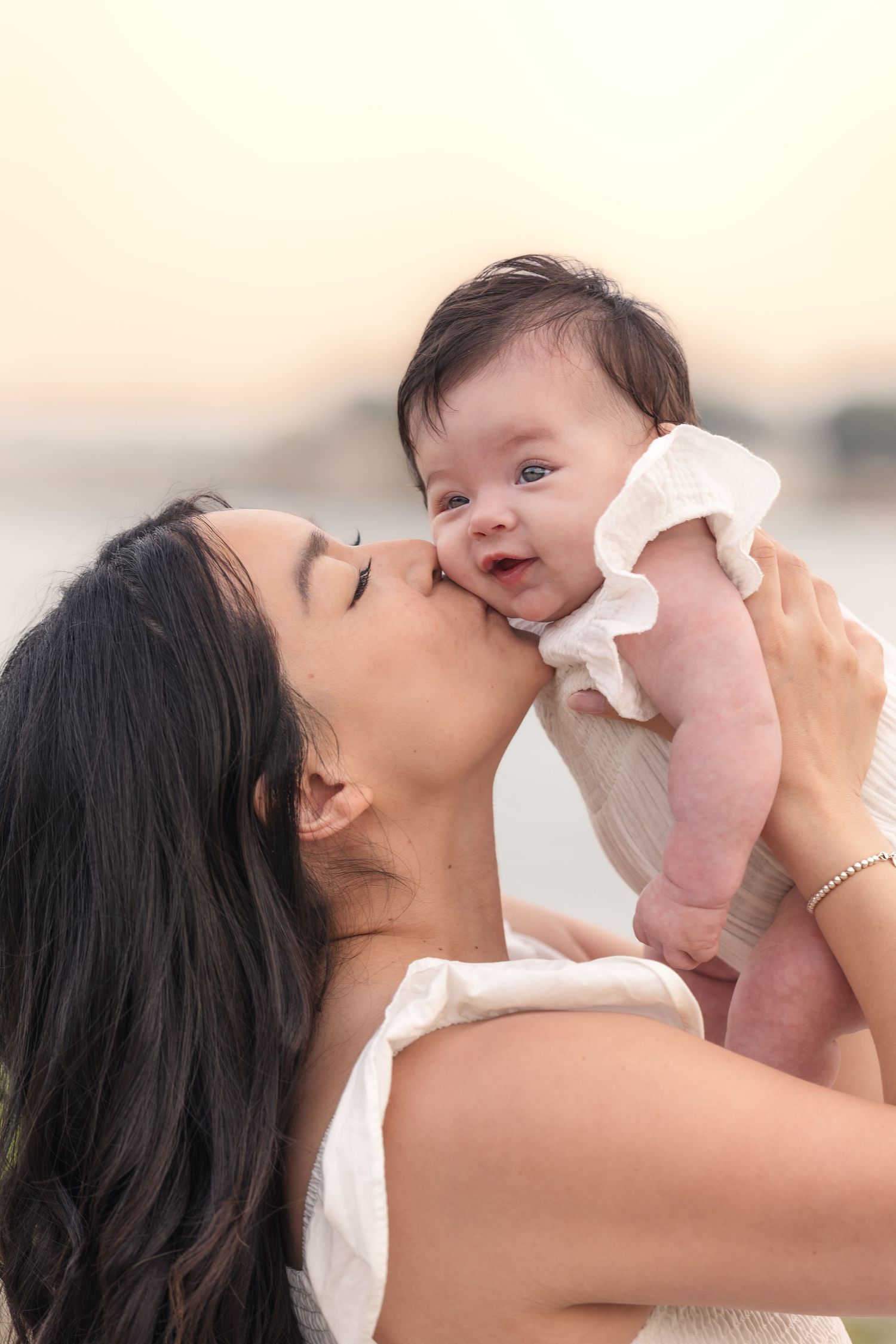 A loving mother kisses her smiling baby girl outdoors at sunset, both wearing white outfits.