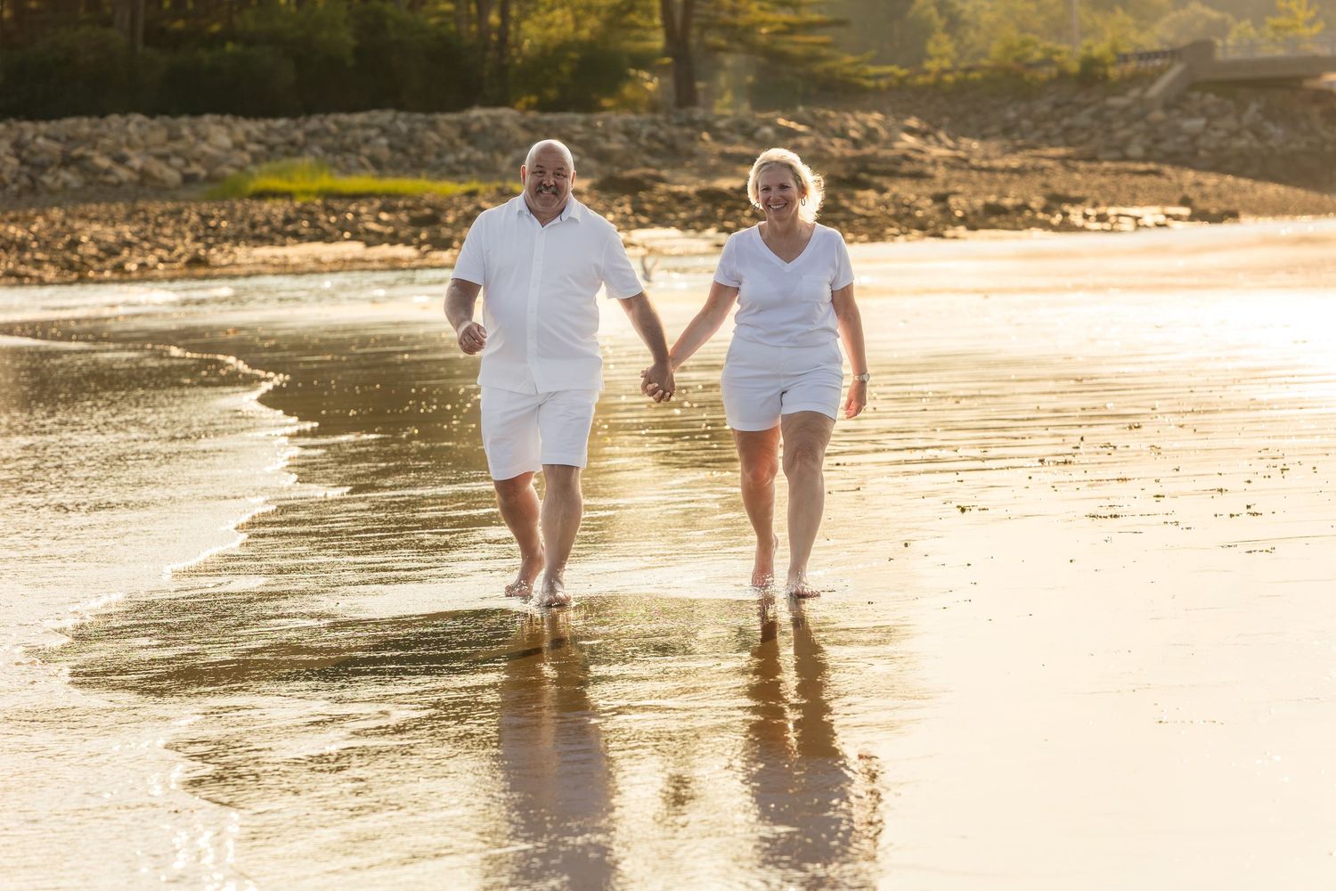 Elderly couple in white outfits holding hands while walking barefoot on a sunny beach shoreline.