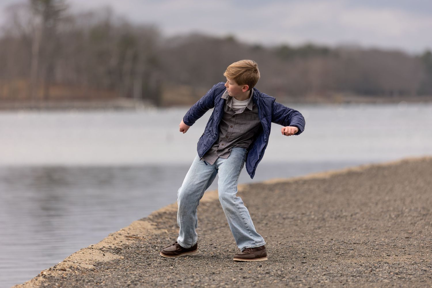 Young boy skipping stones by a calm lake shore on a cloudy day, wearing a blue jacket and jeans.