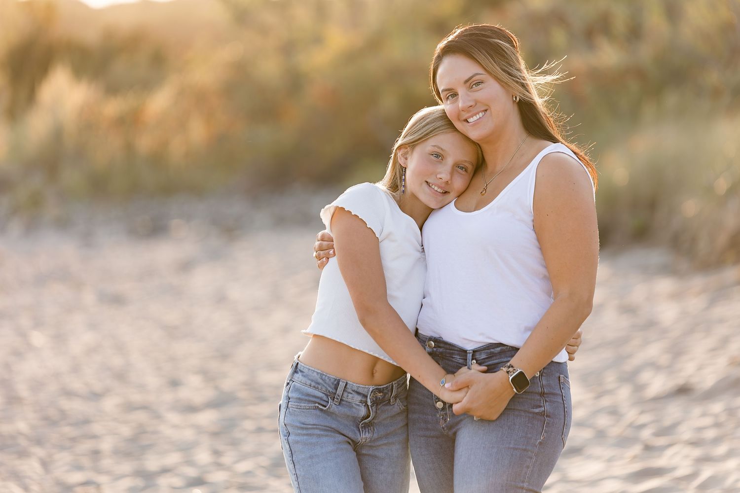 Two women in white tops and jeans embrace warmly on a sandy beach at golden hour.