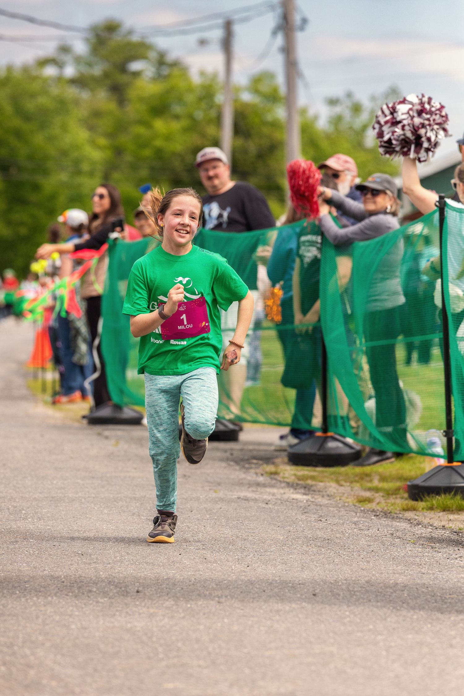 Young girl in green shirt running a race with bib number 1, cheered on by spectators at outdoor event.