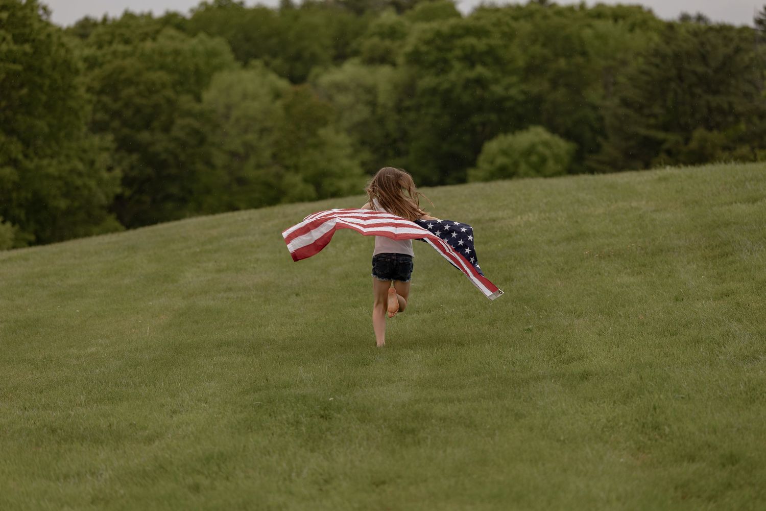 A young girl runs up a green hill with an American flag cape flowing behind her.