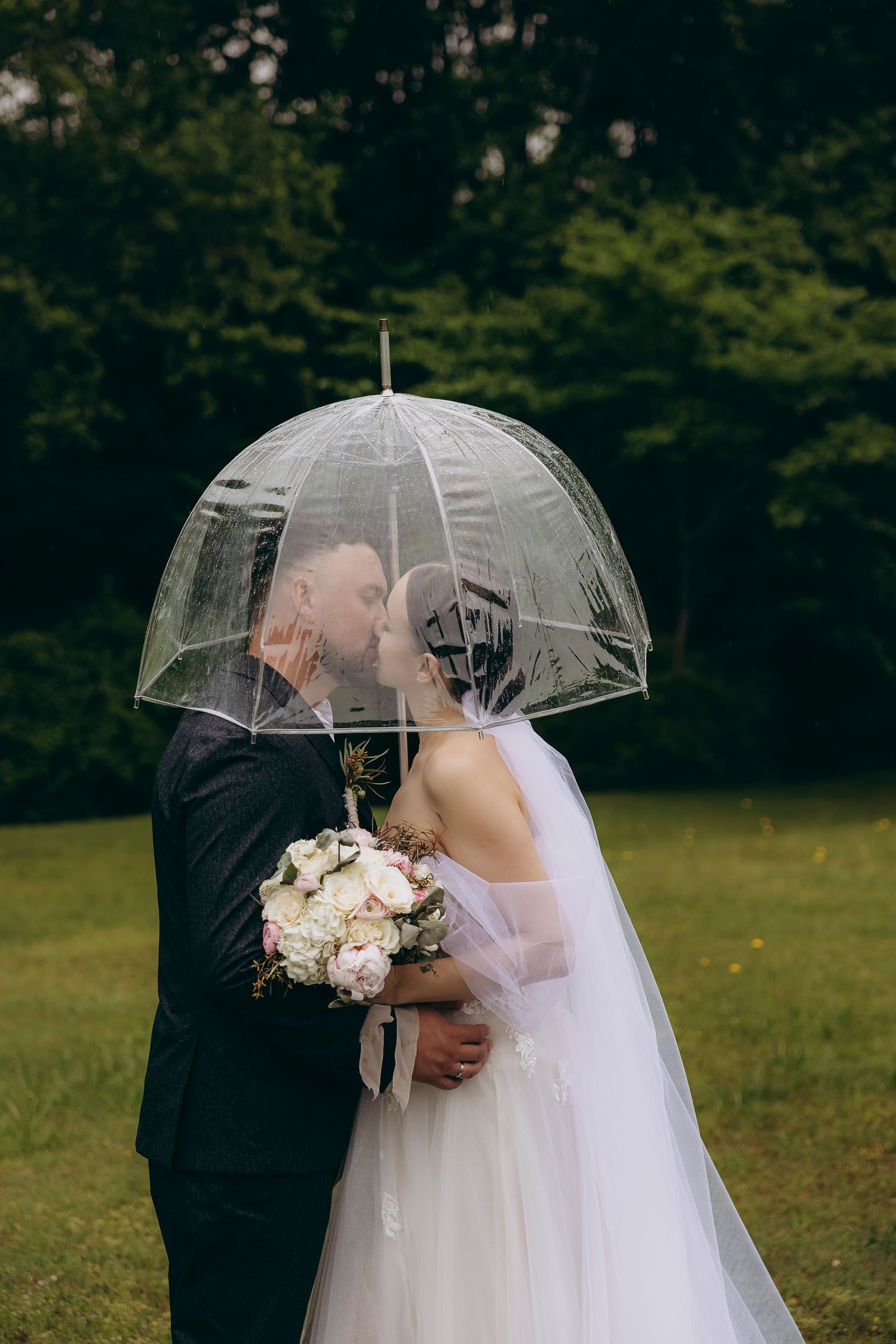 Wedding couple sharing an intimate kiss under a clear bubble umbrella in a green field, Julia Tris Photography.