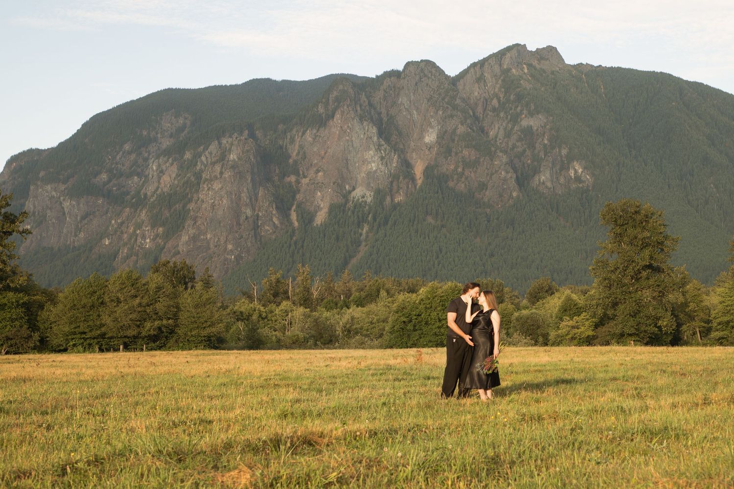 Wide portrait of an expectant couple embracing in a North Bend meadow below Mount Si