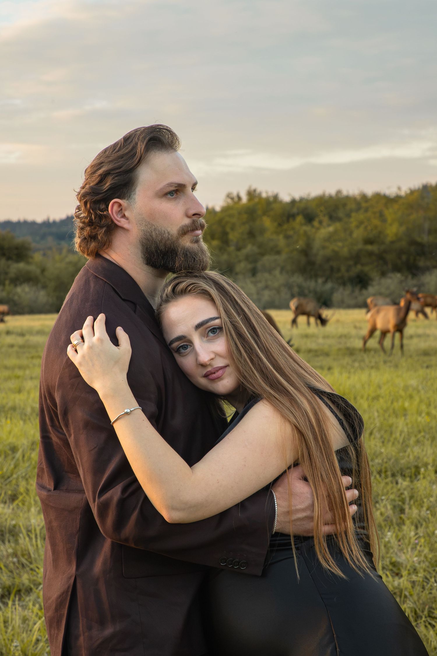 Expectant couple sharing a quiet embrace as a herd of elk grazes behind them in North Bend