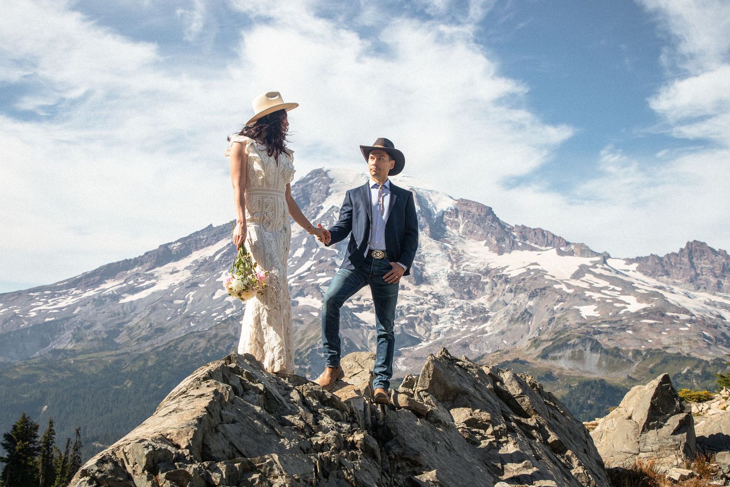 Newlyweds holding hands on a rocky overlook with Mount Rainier behind them