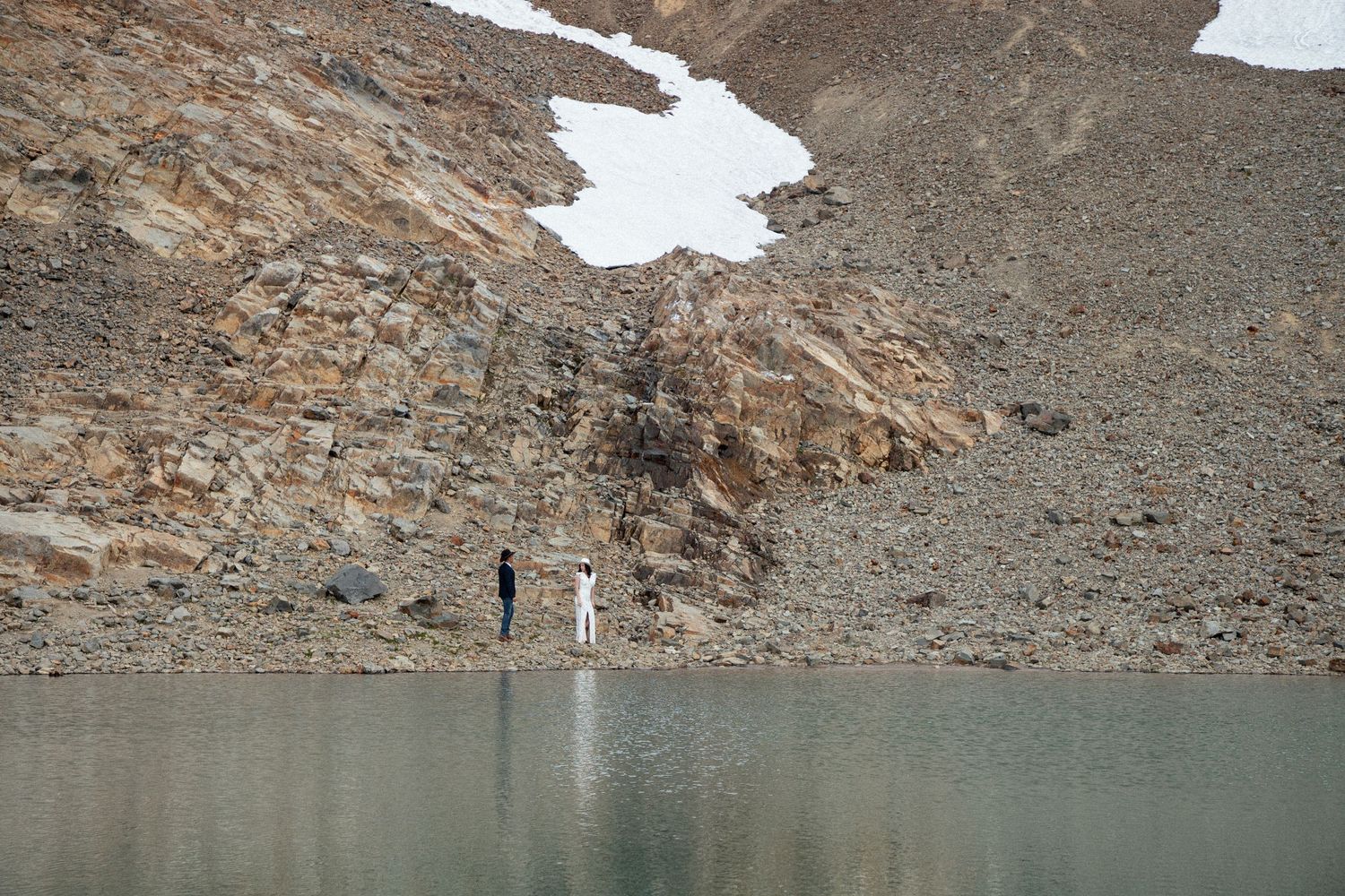 Bride and groom beside a turquoise alpine lake below a rocky, snow-streaked ridge at Mount Rainier