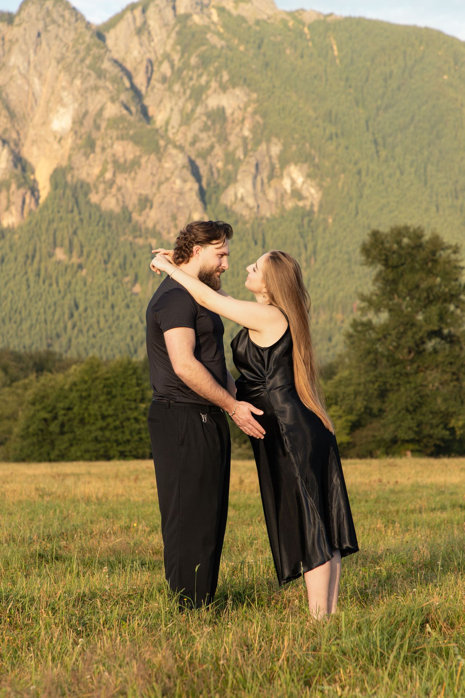 Pregnant woman and her partner holding each other in a field beneath Mount Si