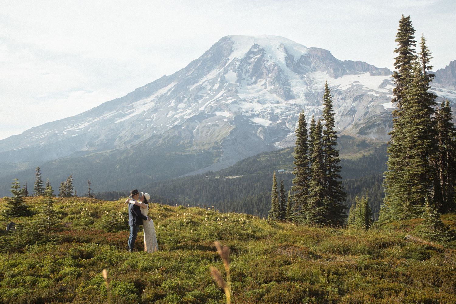 Bride and groom standing in a wild alpine meadow beneath snow-covered Mount Rainier