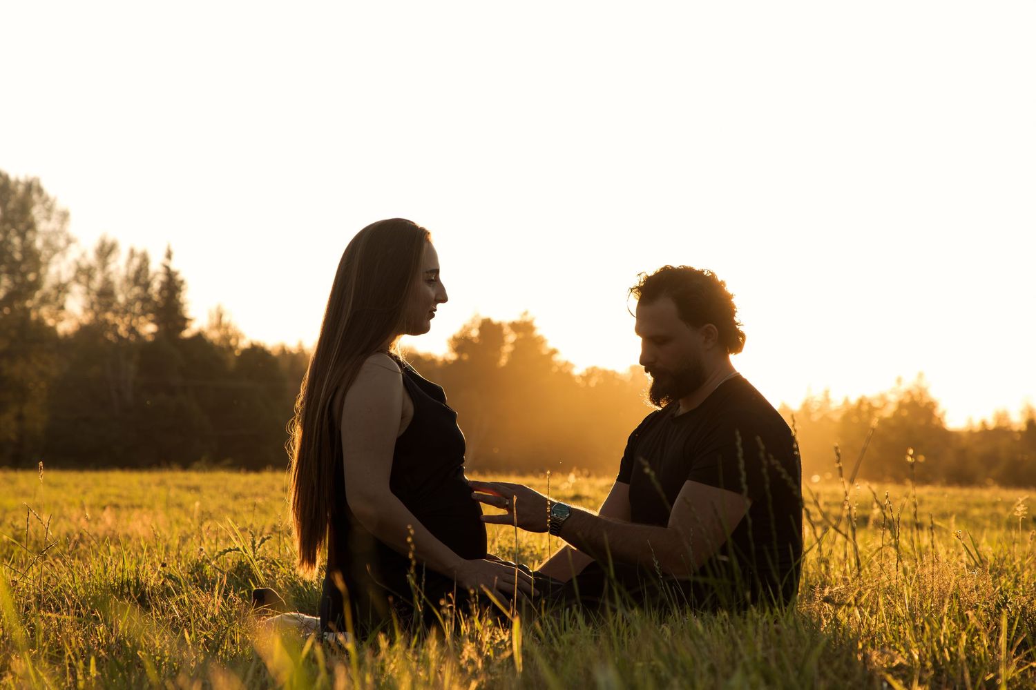 Expectant parents sitting face to face in a sunlit meadow during a romantic maternity photoshoot