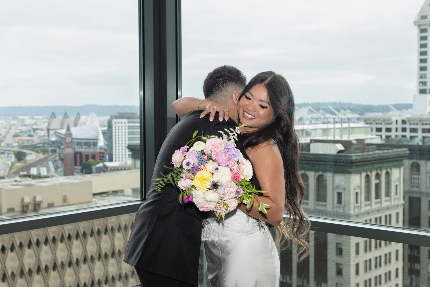 Bride and groom embracing by a large window with downtown views during their Seattle Municipal Court wedding