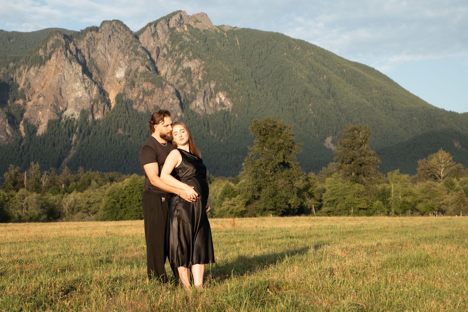 Expectant parents embracing in a meadow with the rocky face of Mount Si behind them