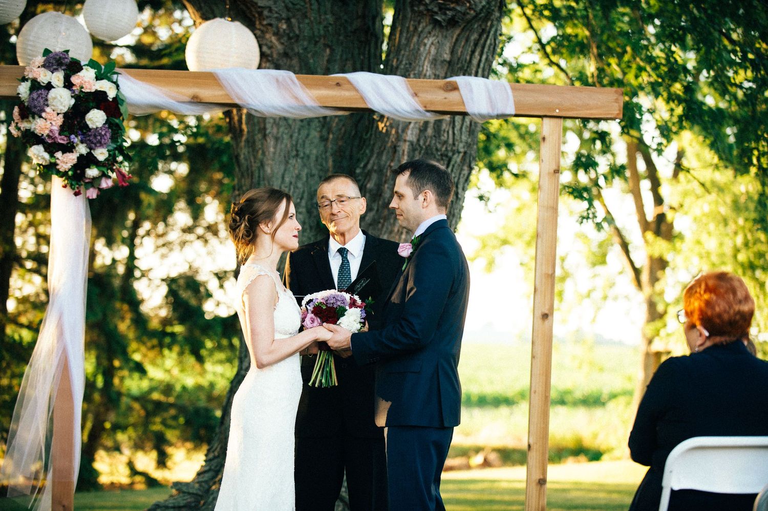 Wedding ceremony setup with white pergola and chairs arranged outdoors among trees.