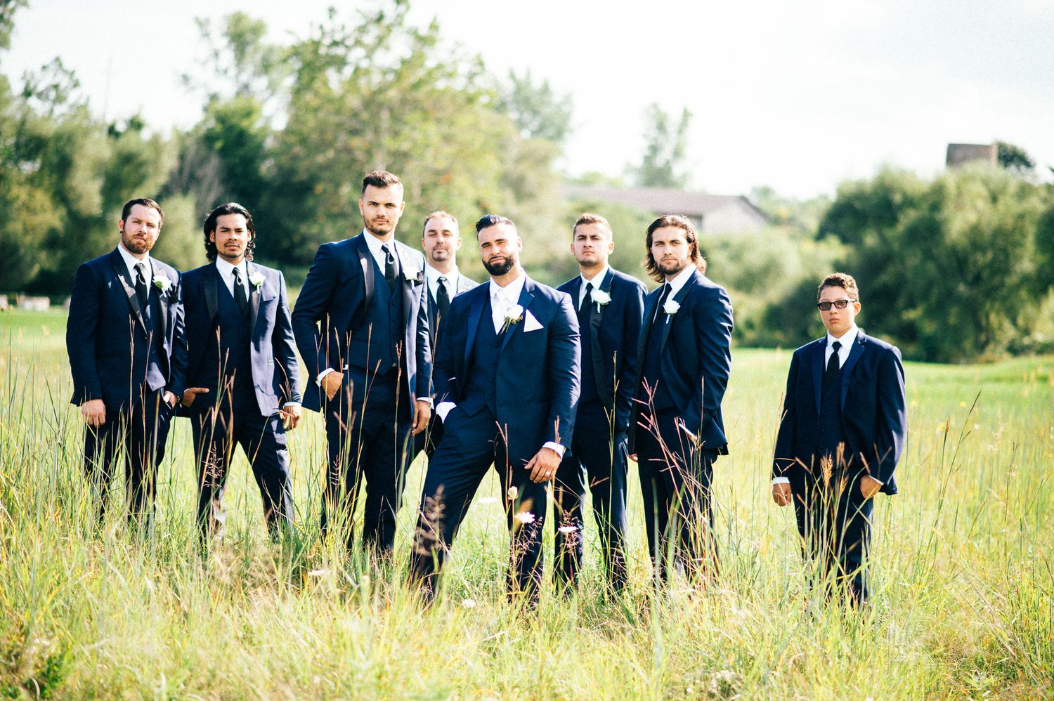 Wedding party in navy suits and white dress stand together in sunny meadow.