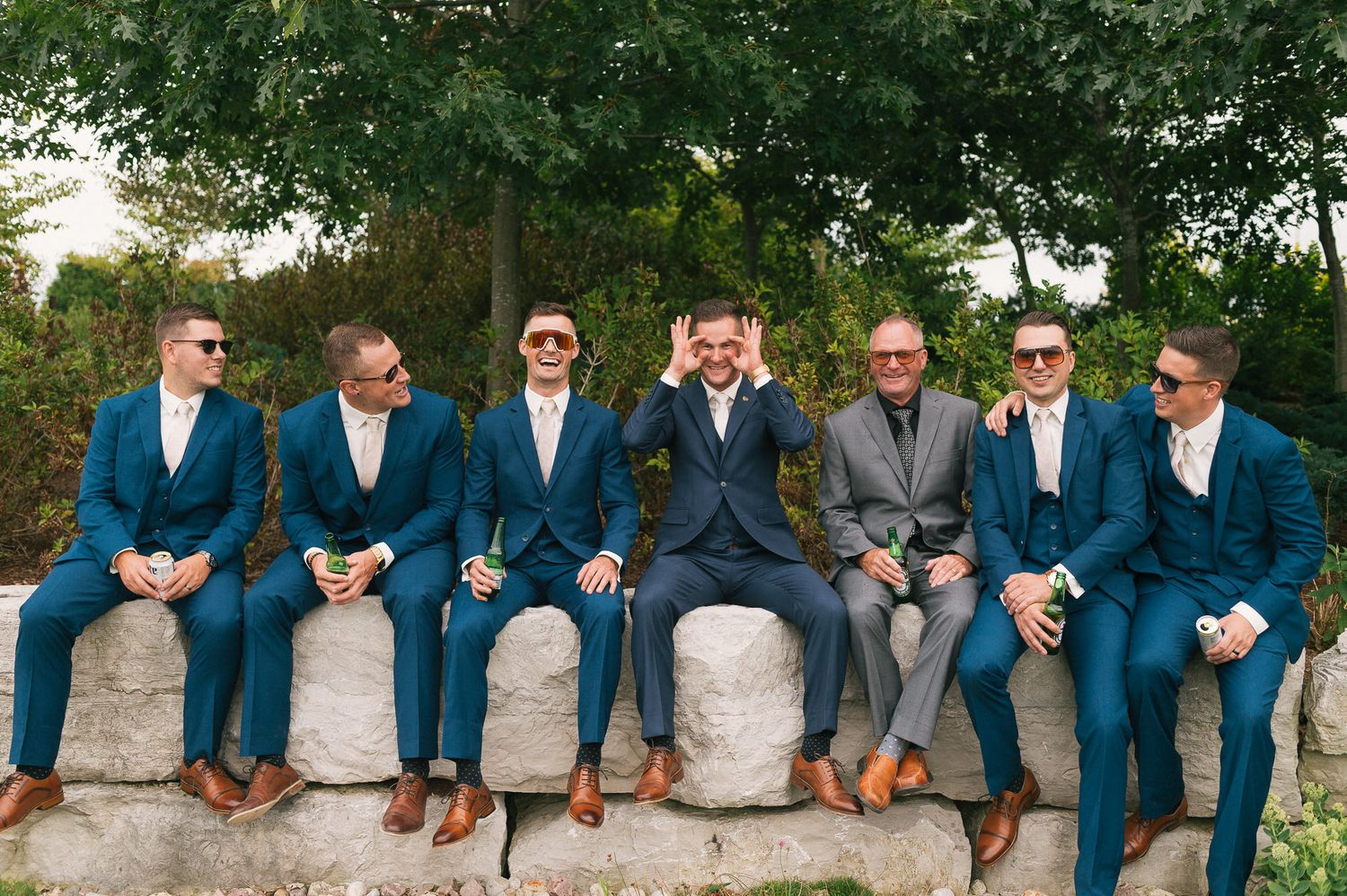 Groomsmen in navy suits sit casually on stone wall showing brown dress shoes.
