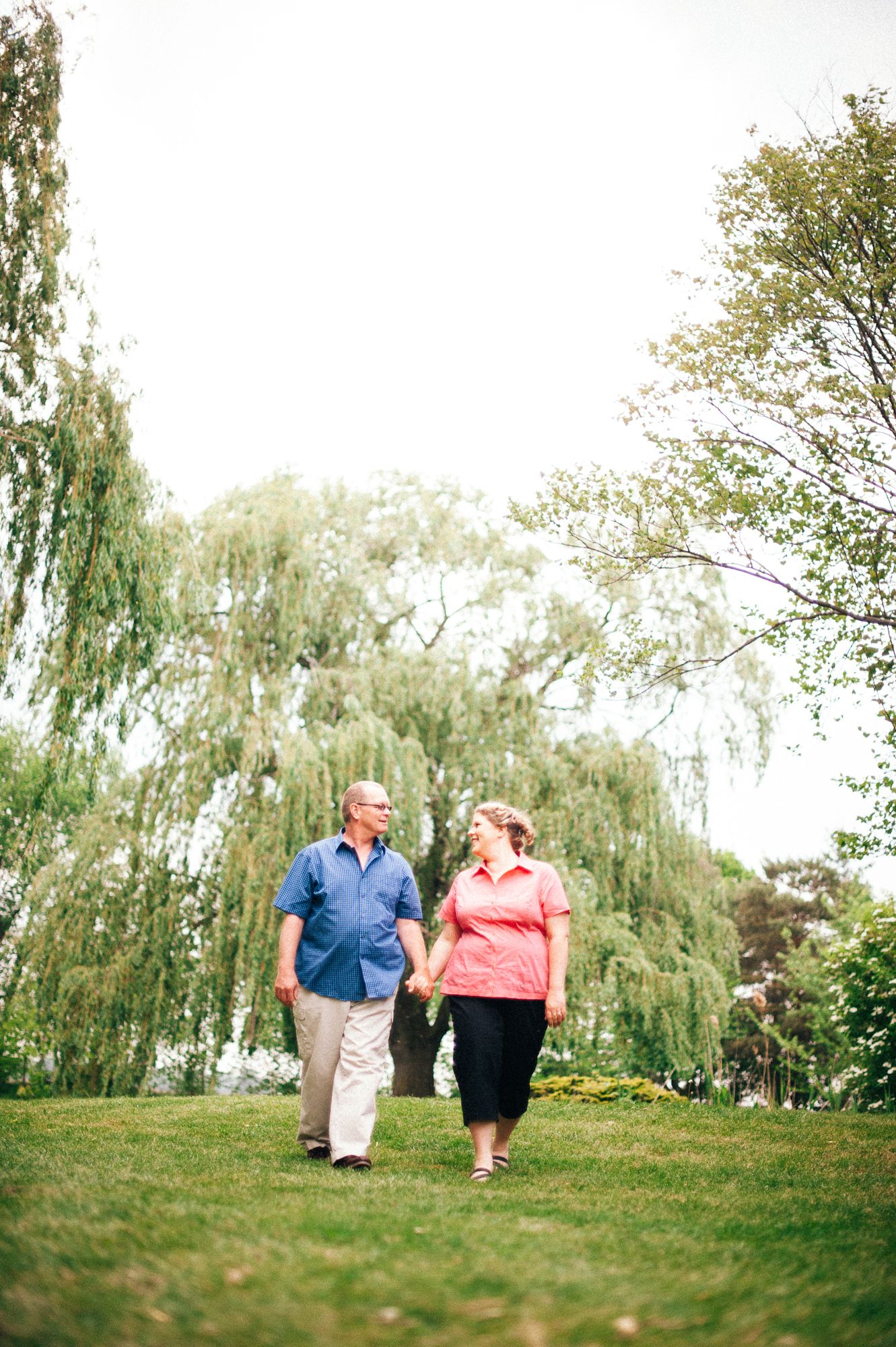 Couple holding hands while walking on grassy hill under weeping willow trees.