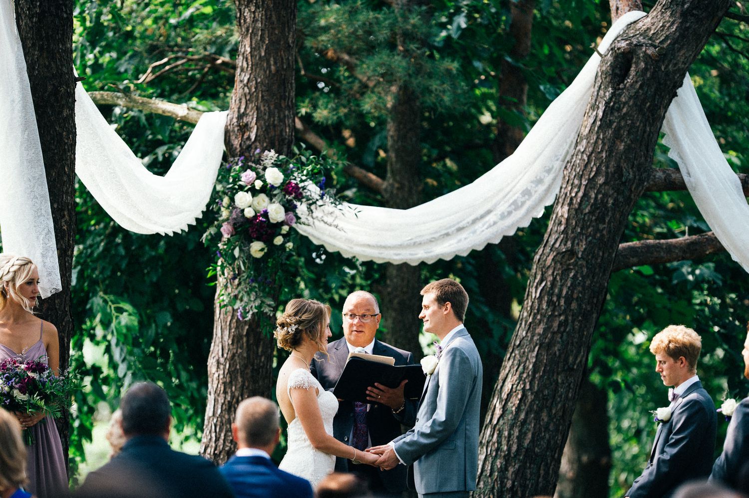 Wedding ceremony taking place outdoors under tall trees with white decorative elements.