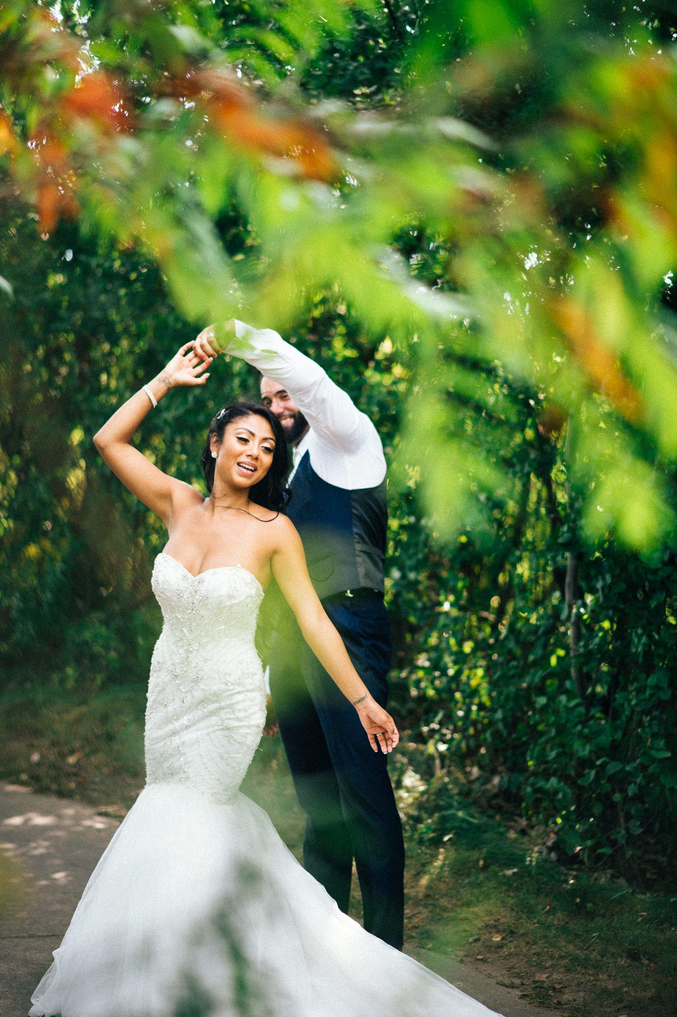 Joyful dancing moment captured through green foliage with flowing white wedding dress.