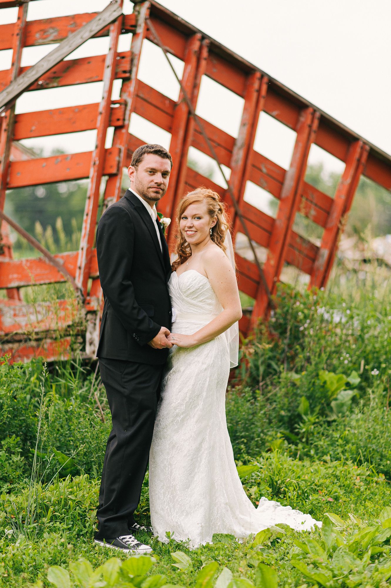 Wedding couple poses playfully by red wooden fencing surrounded by greenery.