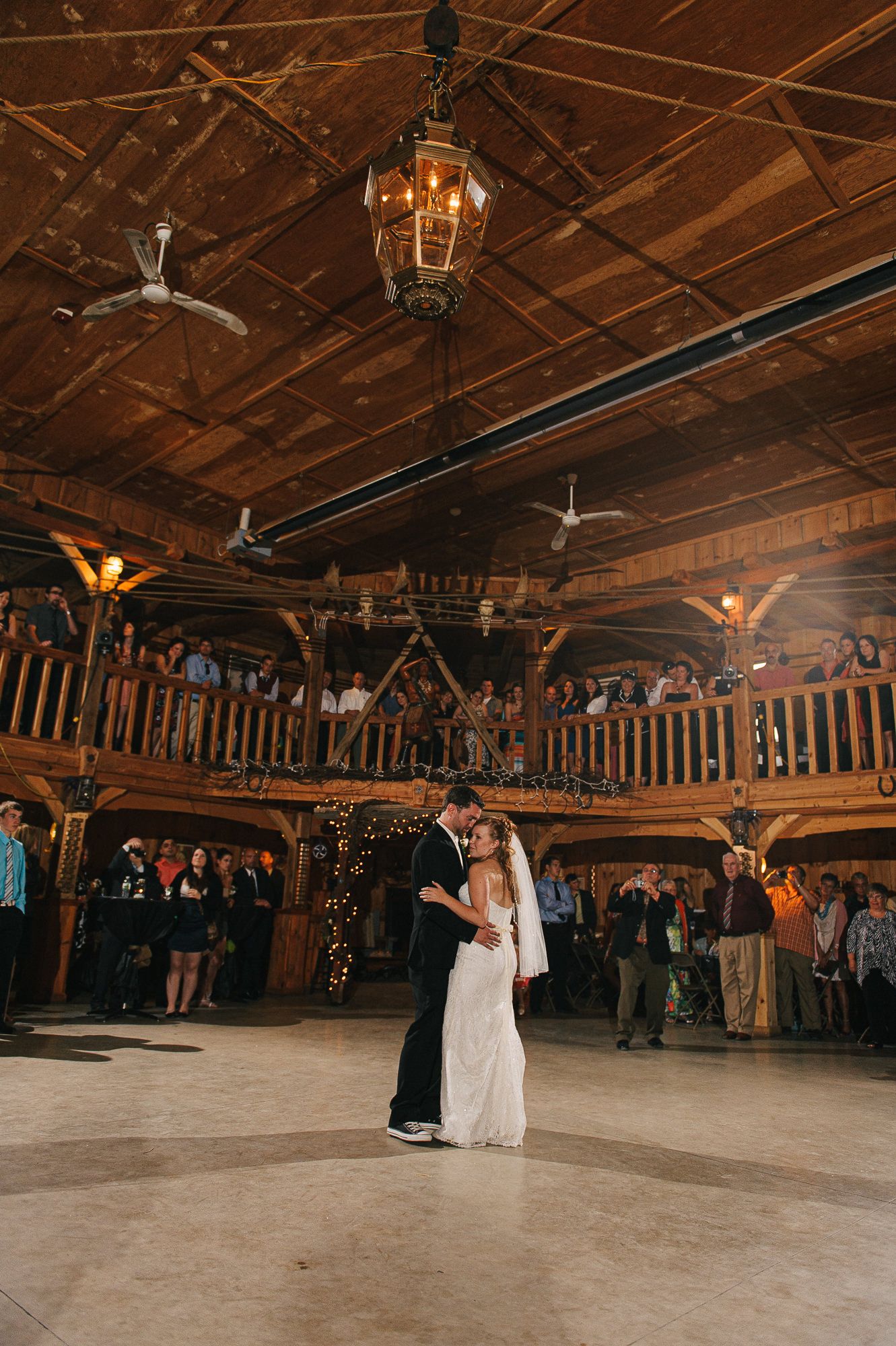 Wedding reception in a rustic wooden barn venue with guests watching from the balcony.