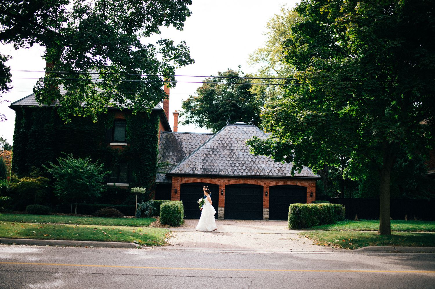 Bride walking near historic brick building surrounded by trees.