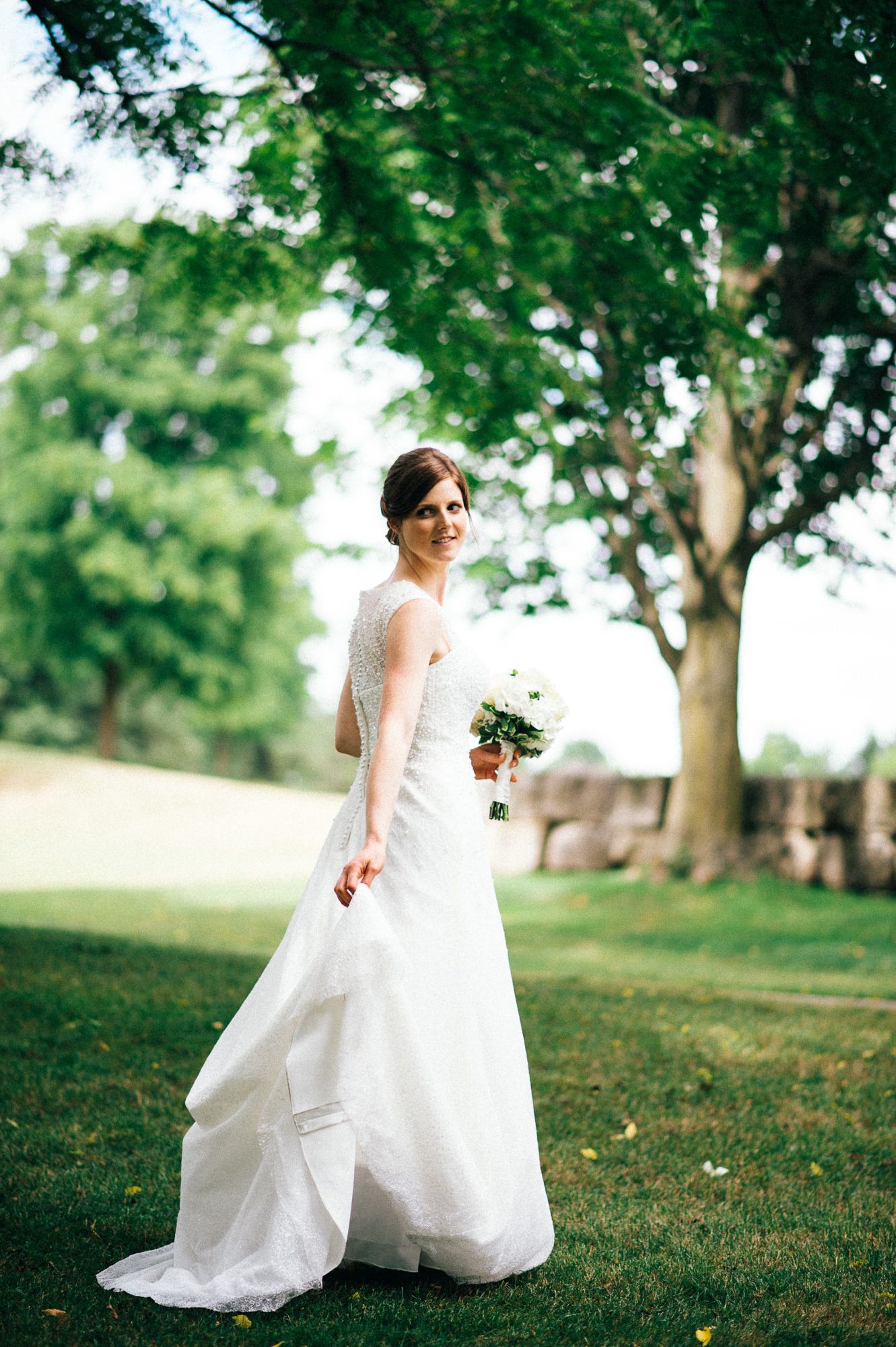 Bride in flowing white gown poses gracefully on grassy lawn with trees in background.