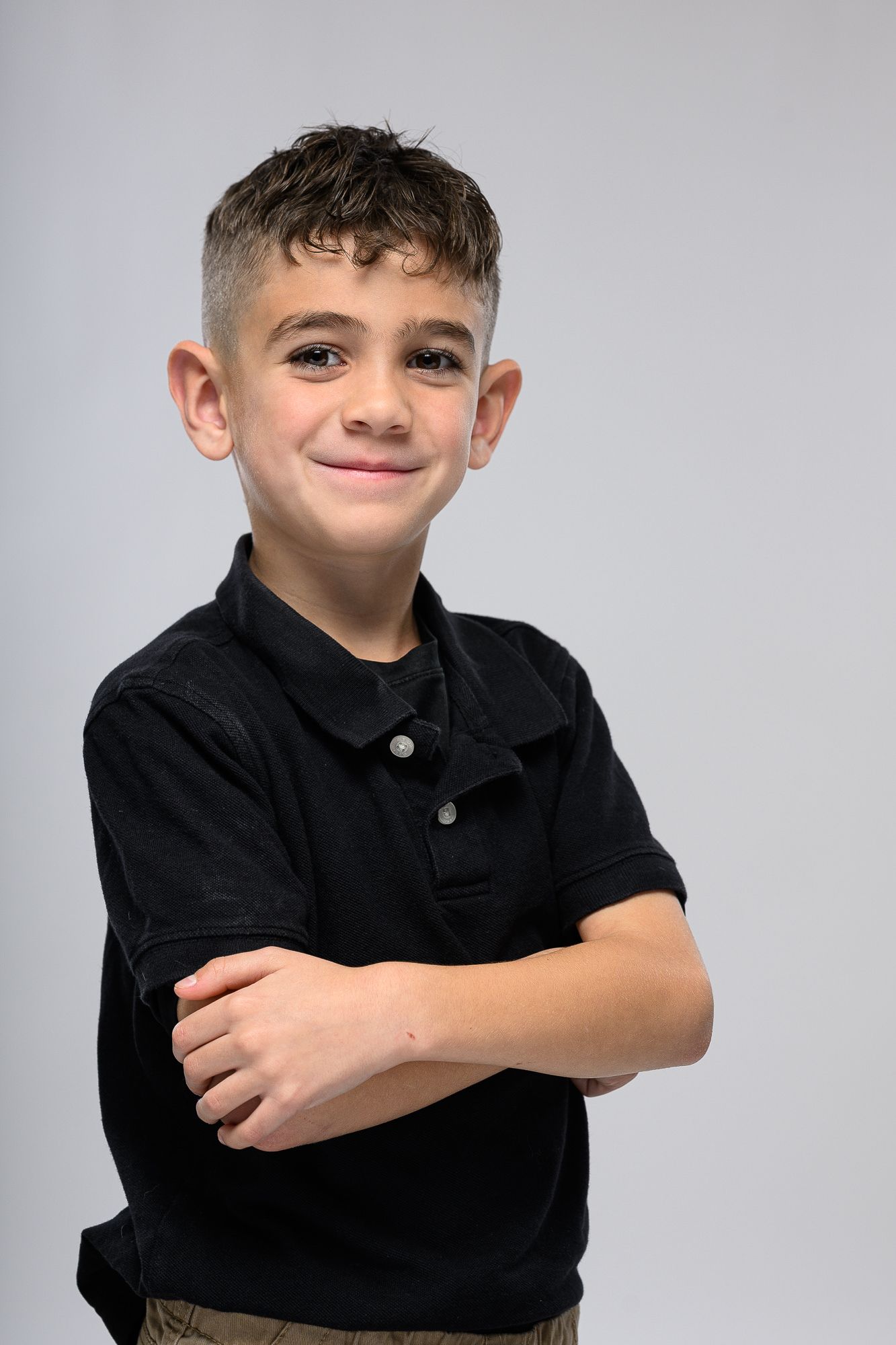 Young child in black polo shirt posing playfully against grey background.