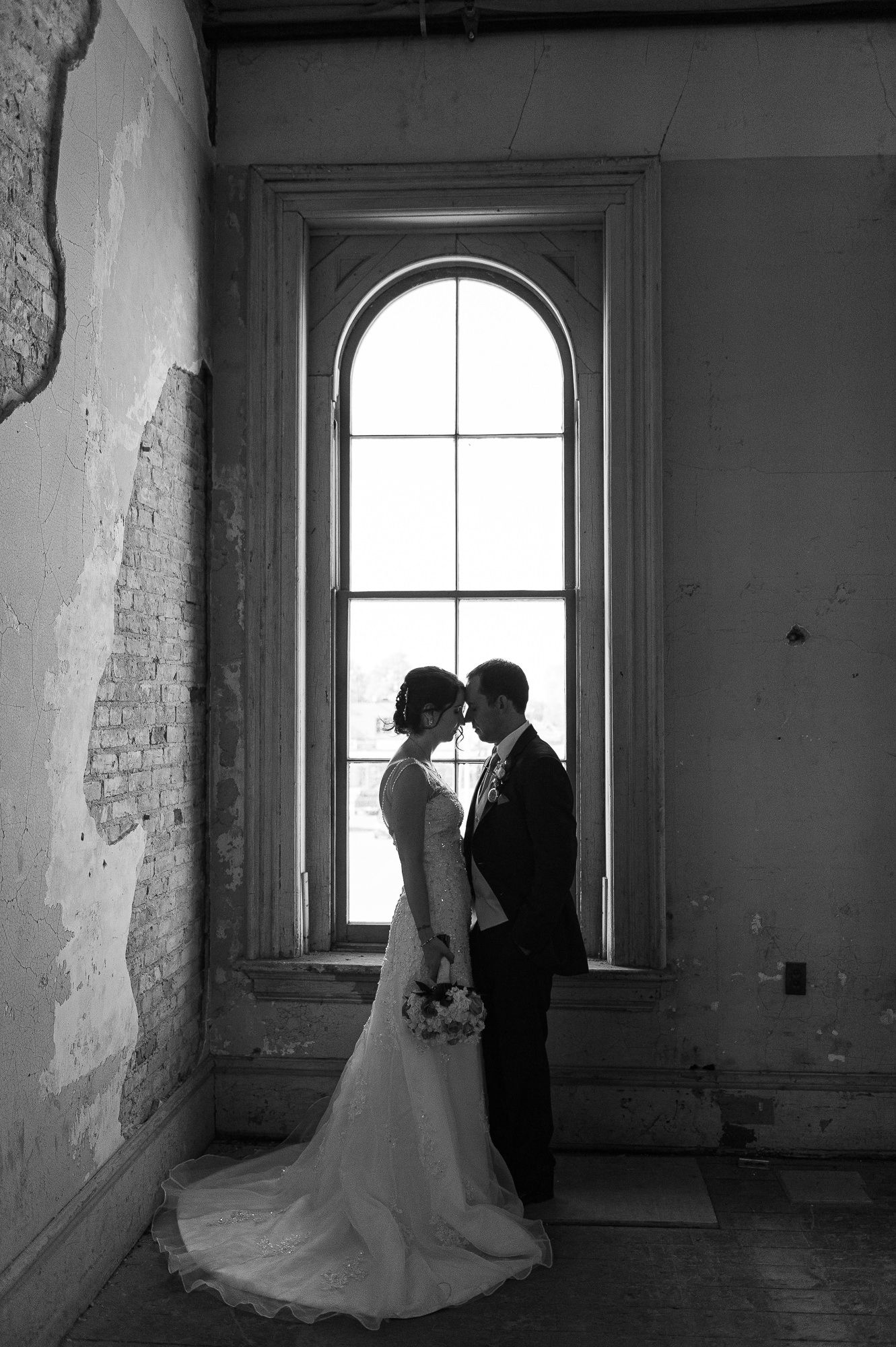 Silhouetted couple in wedding attire by arched windows in rustic interior.