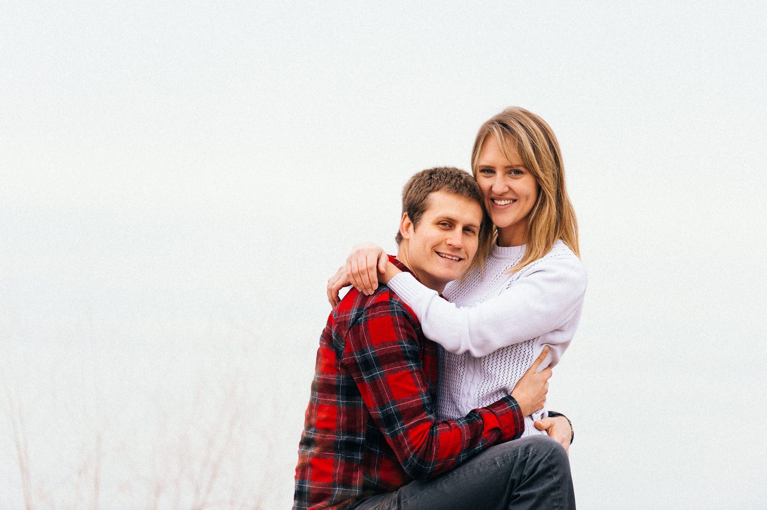 A couple poses together outdoors wearing casual clothing with a red plaid shirt and white top.