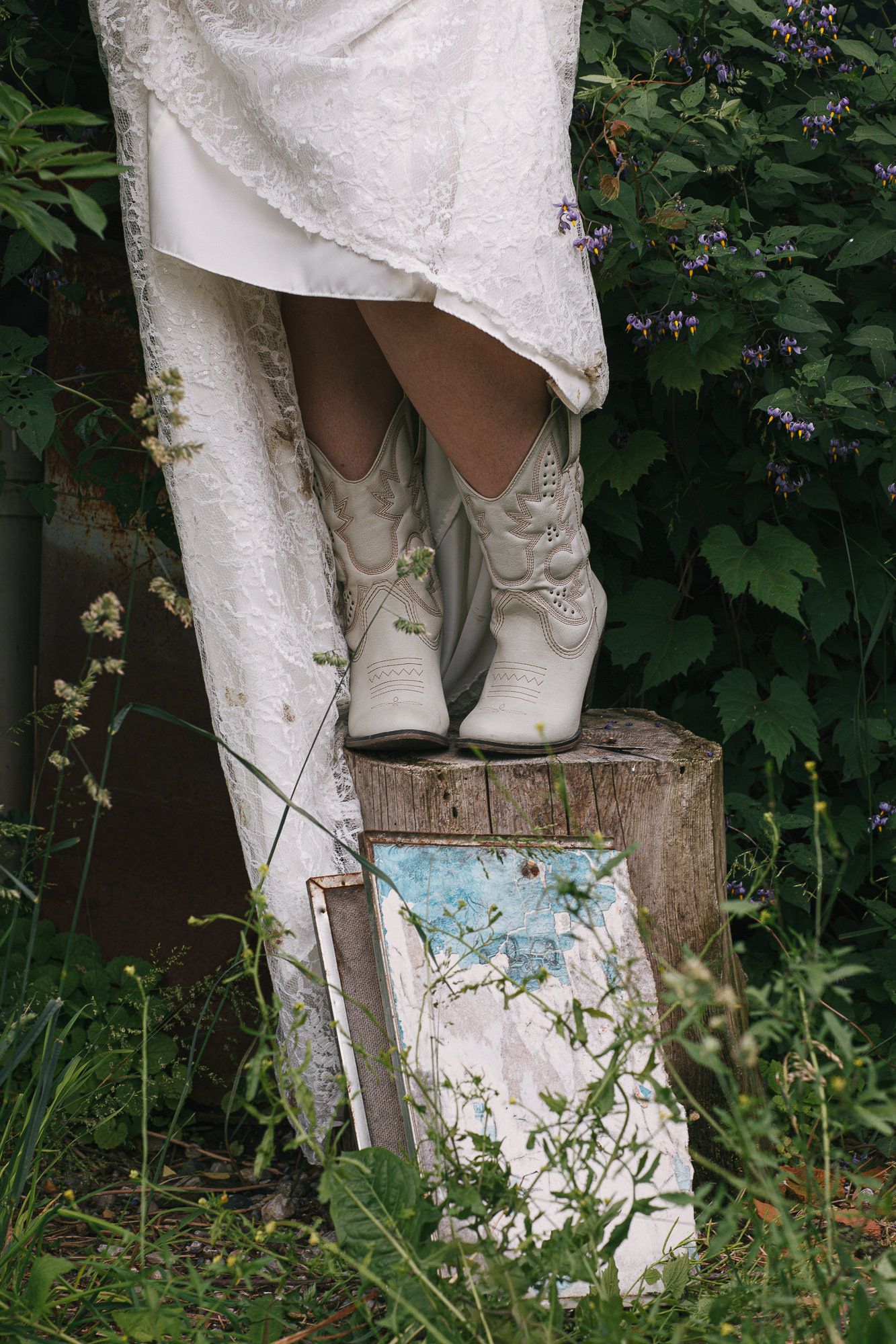 White cowboy boots peek out from under a lace wedding dress against a rustic wooden backdrop.