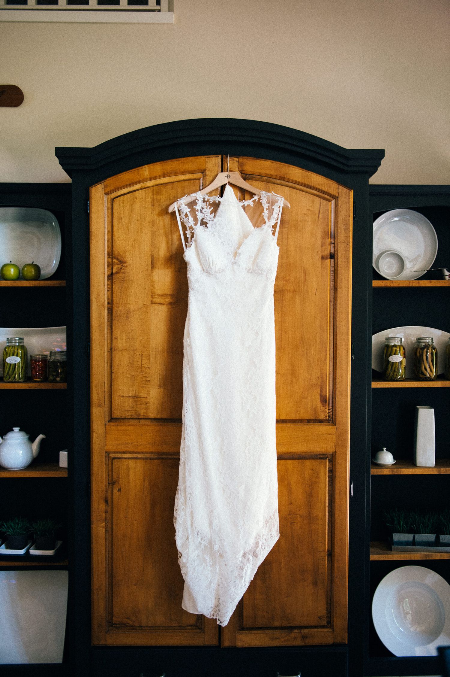 White lace wedding dress hanging on display against wooden armoire and dark walls.