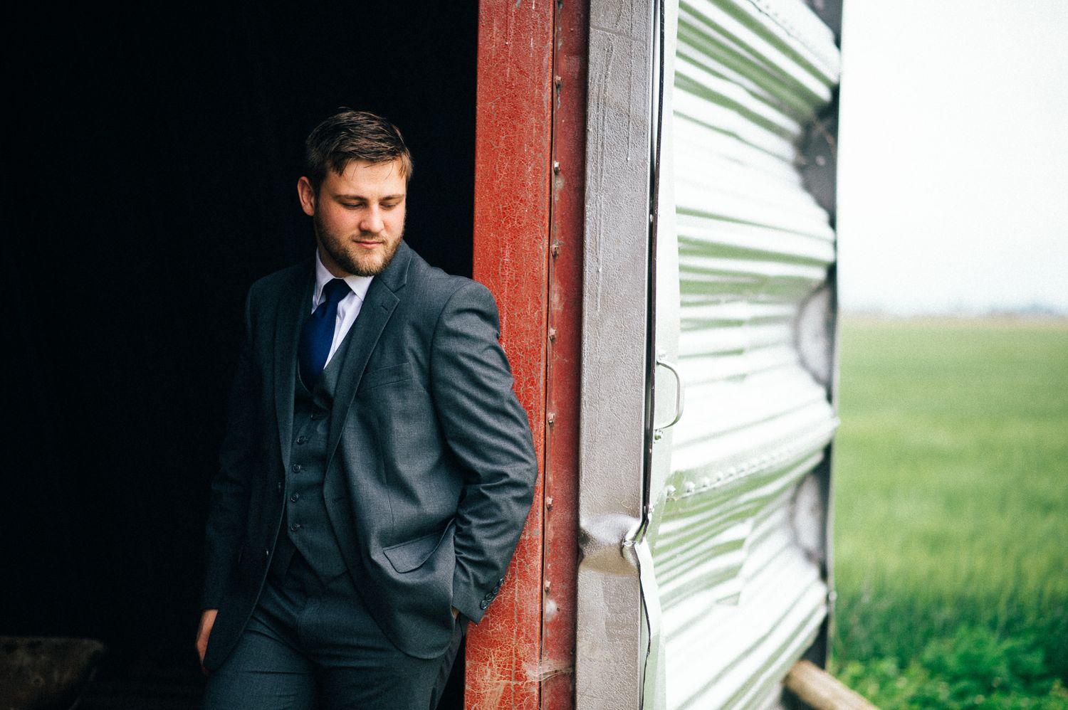 Groom in dark suit leans against rustic red barn wall in countryside setting.