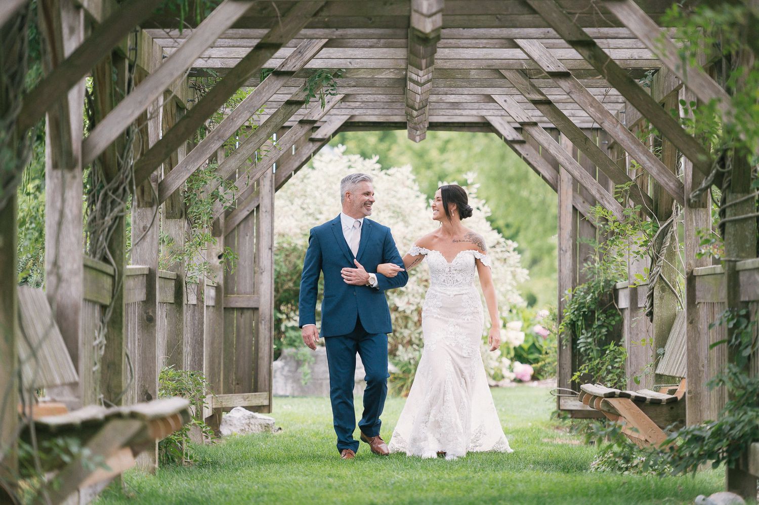 Wedding couple walks through rustic wooden garden arbor covered in climbing vines.