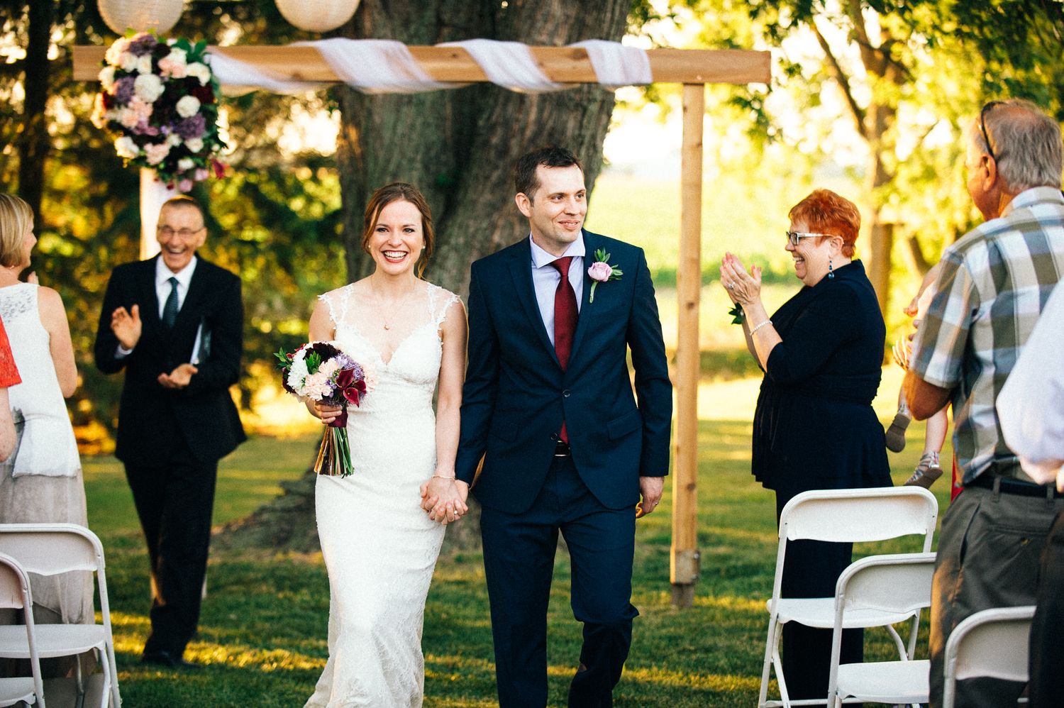 An outdoor wedding ceremony setup with white chairs and guests in a sunlit garden setting.