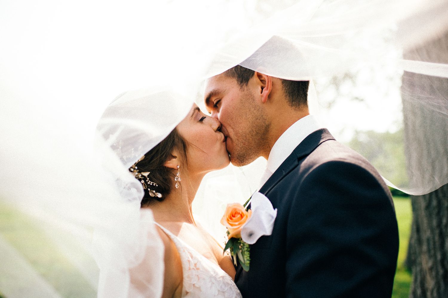 Romantic wedding couple shares an intimate kiss under a flowing white veil in natural light.