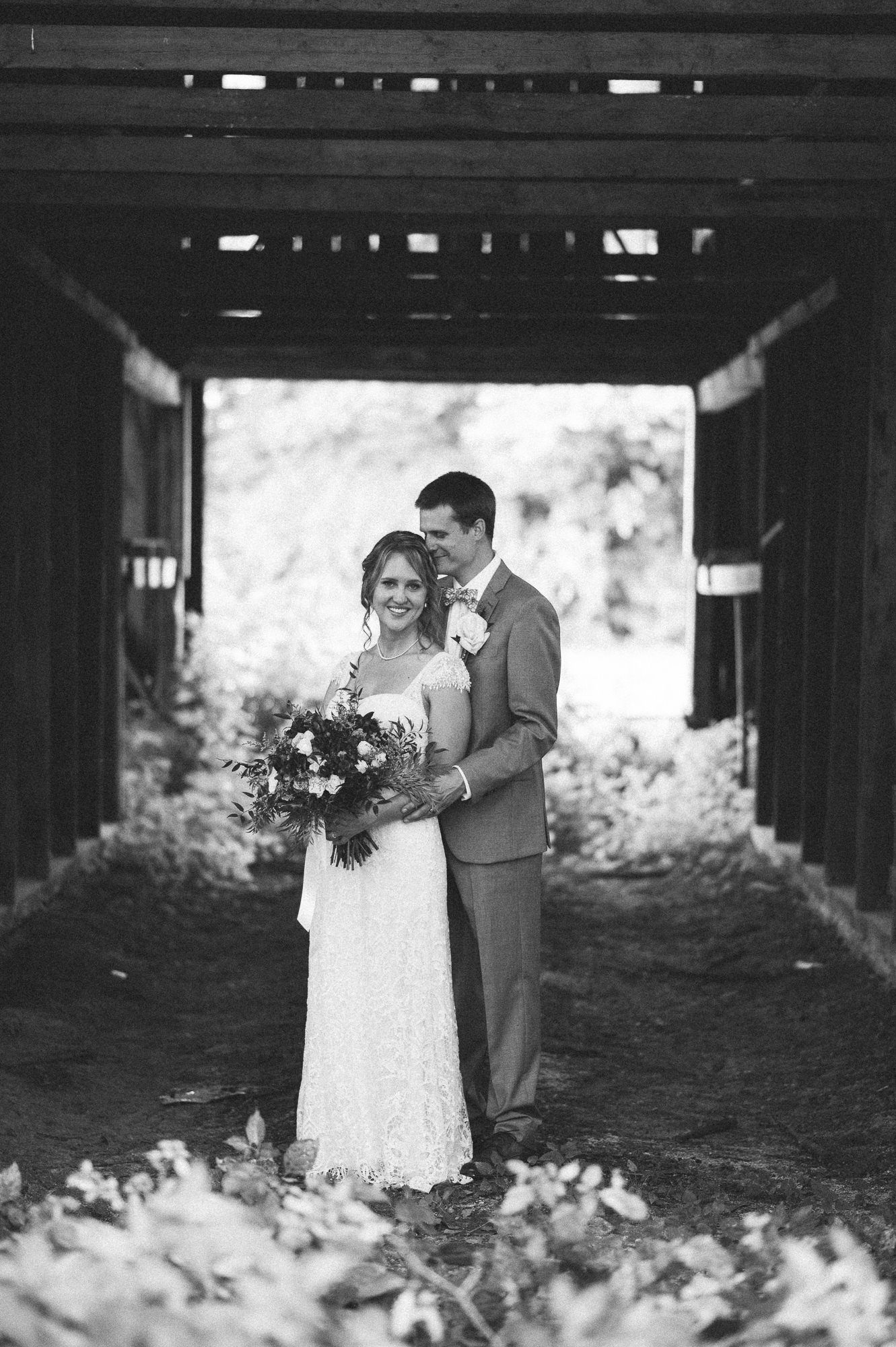 Black and white photo of newlyweds posing in a rustic covered walkway with fallen leaves.