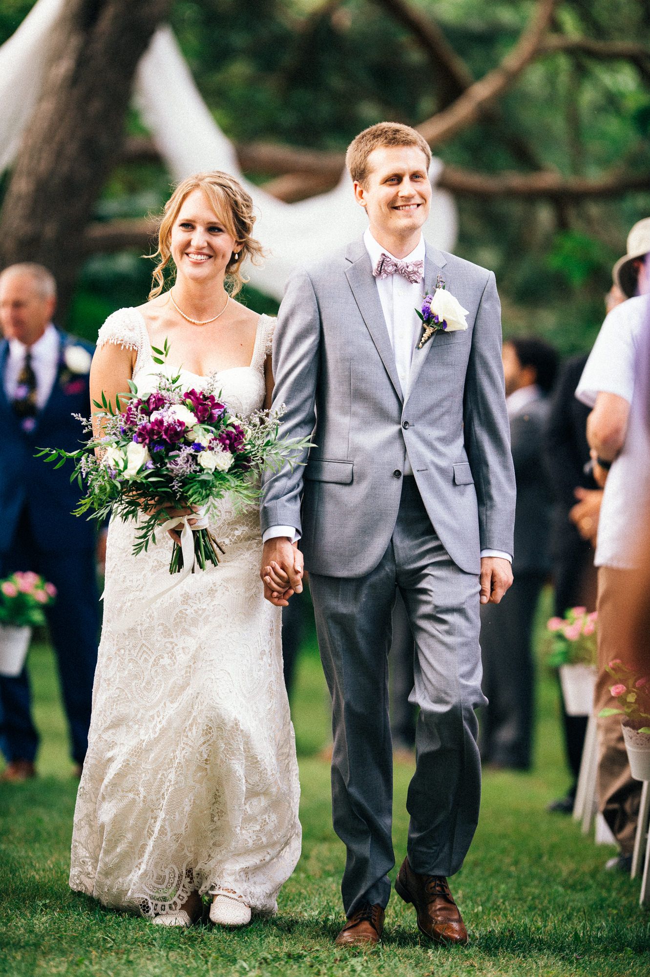 Wedding couple walking together on grass holding beautiful purple bouquet.