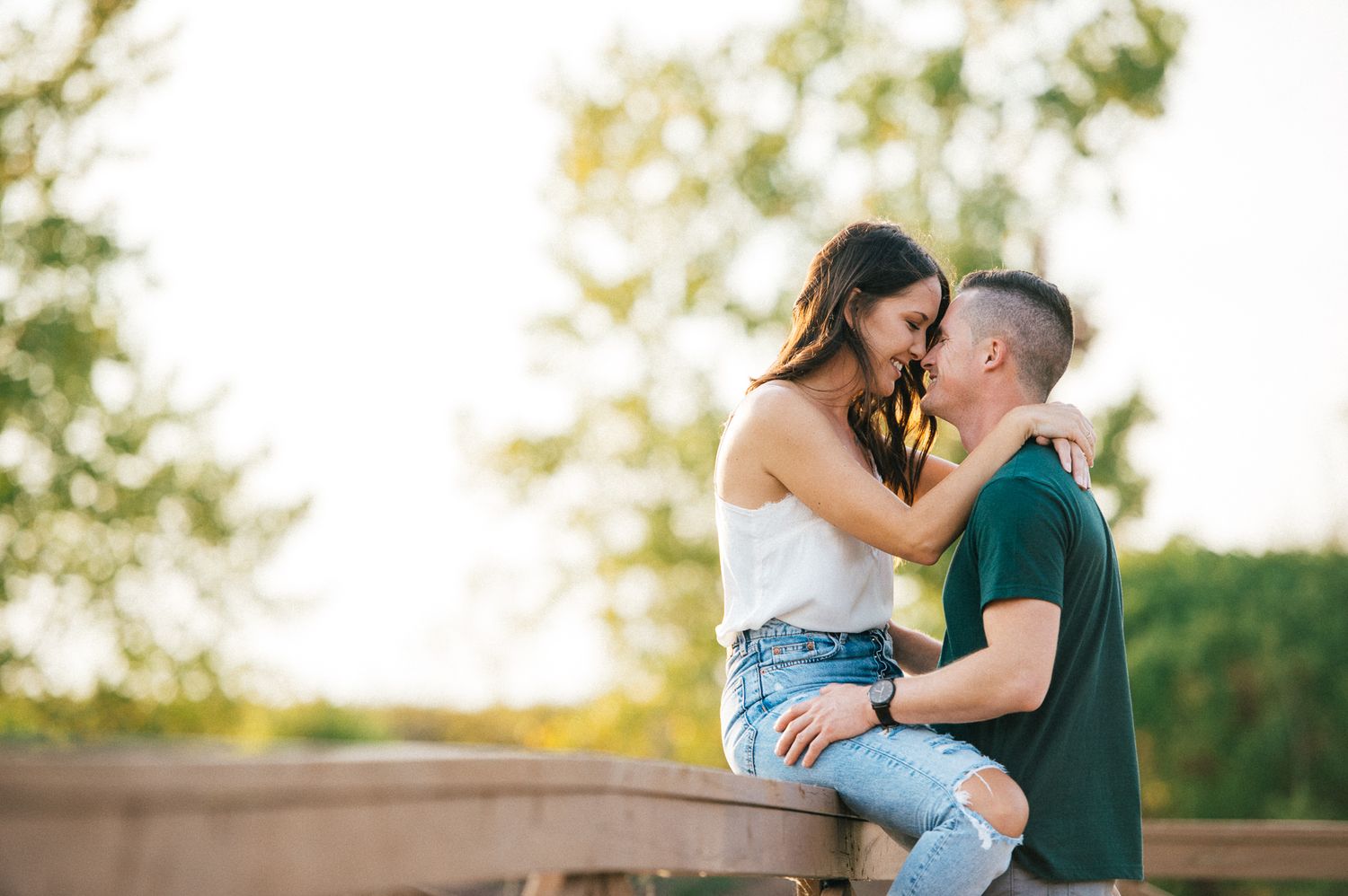 A couple shares an intimate embrace while sitting on a wooden railing at sunset.
