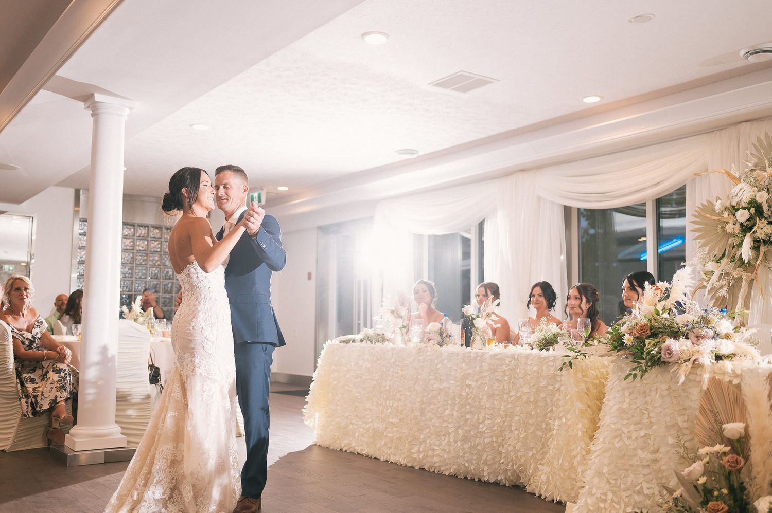 Newlyweds share their first dance in an elegant white reception hall with floral decorations.