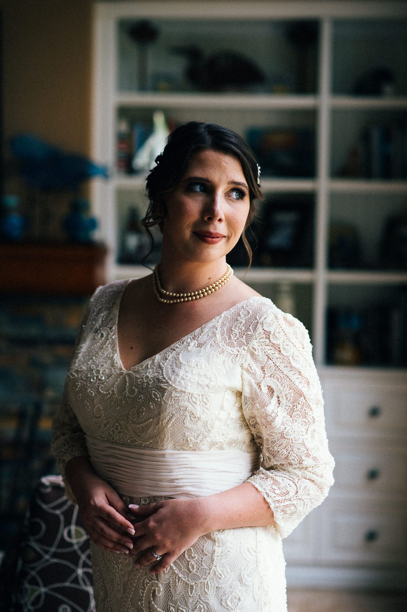 Elegant bridal portrait in white lace gown with pearl necklace against window light.