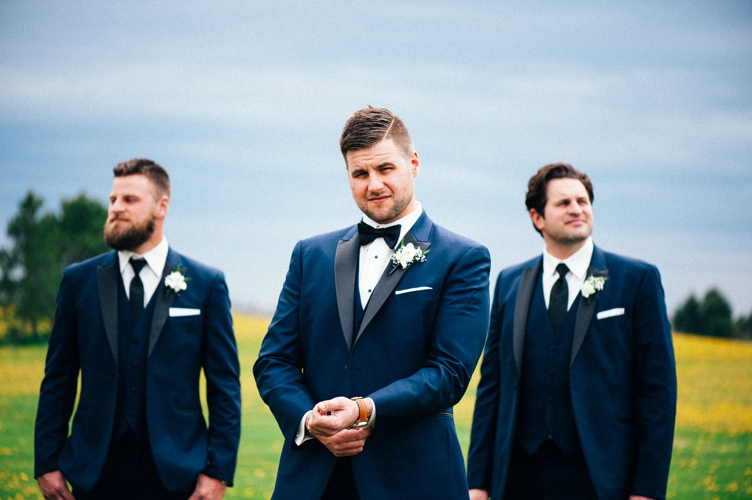 Three groomsmen in navy blue suits stand in a field on a sunny wedding day.